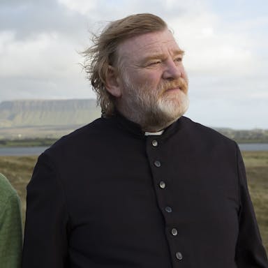 A woman and a man dressed as a priest stand in a windy Irish landscape.