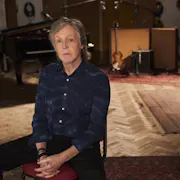 An slim older white man sits in a music studio with a guitar propped up behind him