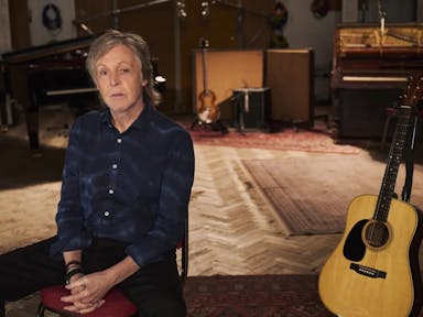 An slim older white man sits in a music studio with a guitar propped up behind him