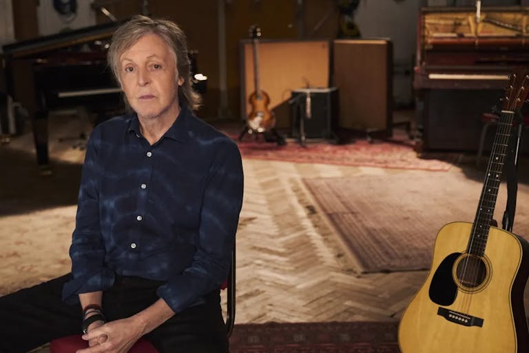 An slim older white man sits in a music studio with a guitar propped up behind him