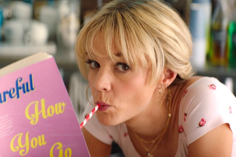 A blonde white woman drinking through a red and white striped straw reading a book with baby pink cover