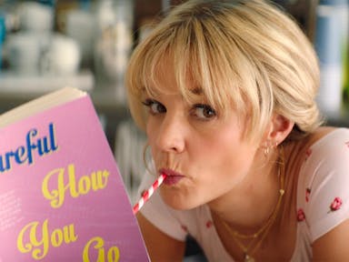 A blonde white woman drinking through a red and white striped straw reading a book with baby pink cover