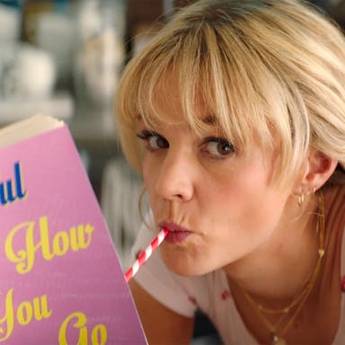 A blonde white woman drinking through a red and white striped straw reading a book with baby pink cover