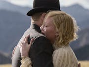Woman with short blonde hair embraces a man in a bowler hat against a backdrop of mountains.
