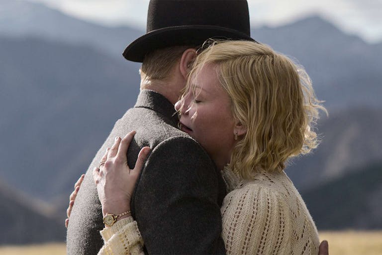 Woman with short blonde hair embraces a man in a bowler hat against a backdrop of mountains. 