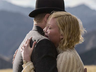 Woman with short blonde hair embraces a man in a bowler hat against a backdrop of mountains. 