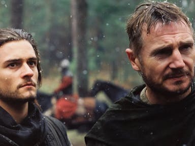 Two white men stand in a slightly snowy battle field, one holding a sword