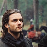 Two white men stand in a slightly snowy battle field, one holding a sword