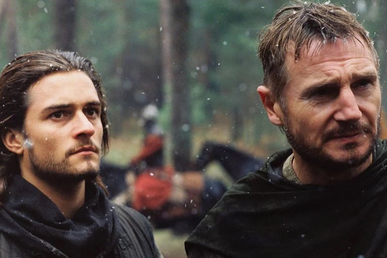 Two white men stand in a slightly snowy battle field, one holding a sword