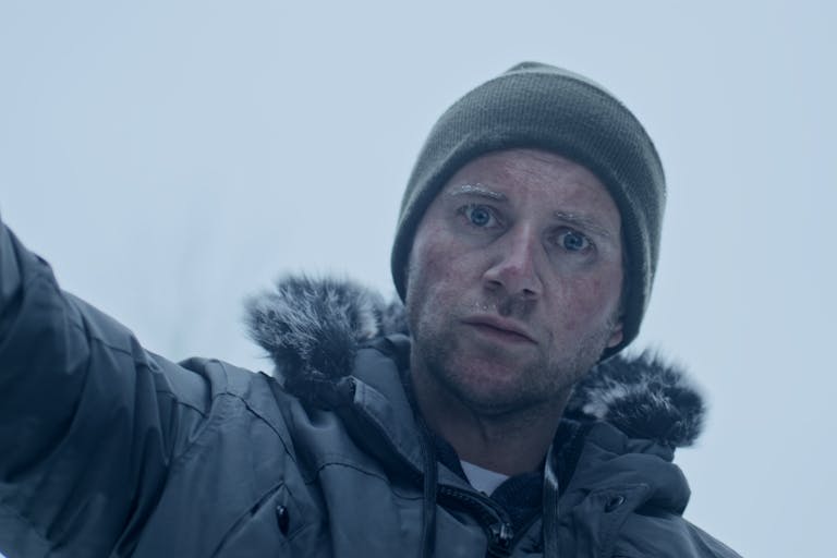 A white man in a freezing, snowy setting, with a woollen beanie hat and coat with fur trim, looking cold and intense 