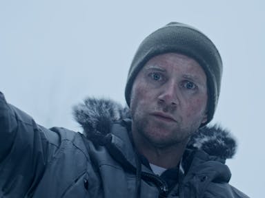 A white man in a freezing, snowy setting, with a woollen beanie hat and coat with fur trim, looking cold and intense 