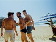 Two white men in speedos walking on a sunny beach with their arms around each other, confronted by another white man