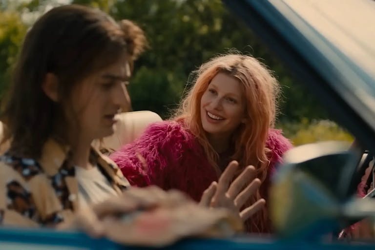 A young white woman with pastel pink hair and hot pink fluffy jacket smiling at a young white man with long brown hair looking stressed, sitting in an open top car
