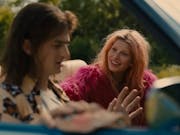 A young white woman with pastel pink hair and hot pink fluffy jacket smiling at a young white man with long brown hair looking stressed, sitting in an open top car