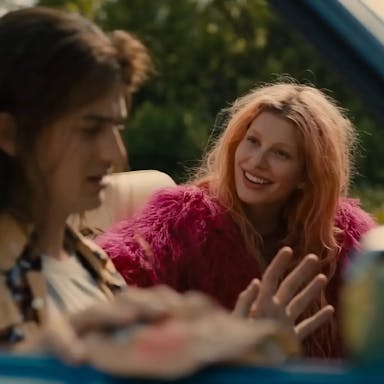 A young white woman with pastel pink hair and hot pink fluffy jacket smiling at a young white man with long brown hair looking stressed, sitting in an open top car