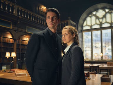 A white man and woman in dark suits stand in a grand library 