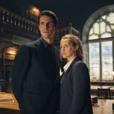 A white man and woman in dark suits stand in a grand library 