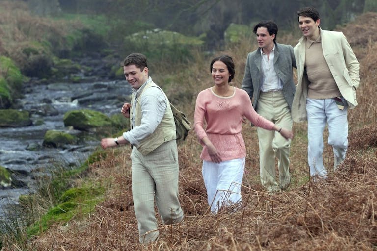 Three young men in light coloured suits and a young woman in a pink top run down a hill next to a stream in the countryside