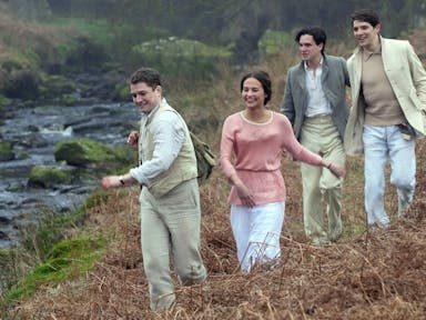 Three young men in light coloured suits and a young woman in a pink top run down a hill next to a stream in the countryside