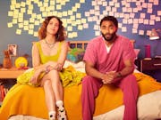 A young white woman in a yellow dress and a young Black man in pink scrubs sitting on a bed with yellow and pink post-it notes behind them on the wall