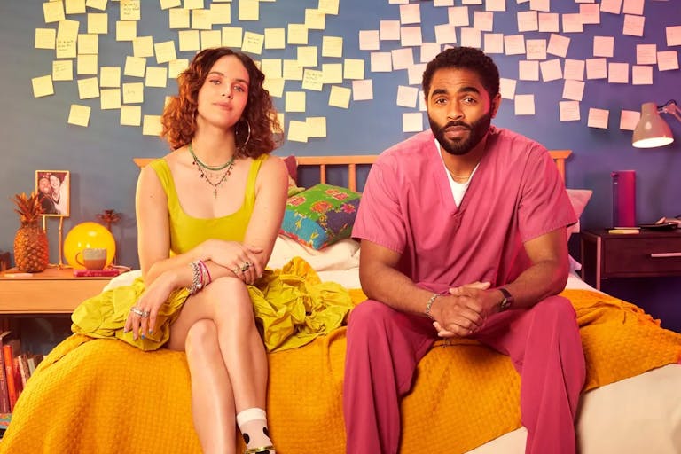 A young white woman in a yellow dress and a young Black man in pink scrubs sitting on a bed with yellow and pink post-it notes behind them on the wall