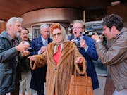 A middle aged white woman with red hair in an updo and silk hair wrap, wearing a fur coat with a cigarette in her mouth, being surrounded by photographers and reports