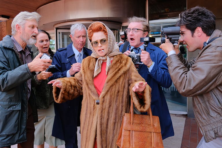 A middle aged white woman with red hair in an updo and silk hair wrap, wearing a fur coat with a cigarette in her mouth, being surrounded by photographers and reports