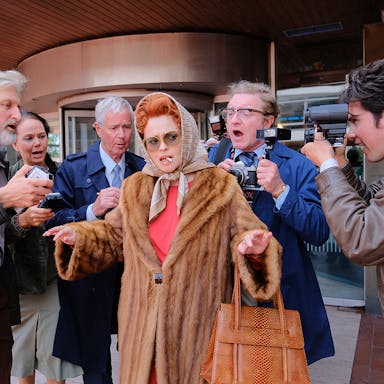 A middle aged white woman with red hair in an updo and silk hair wrap, wearing a fur coat with a cigarette in her mouth, being surrounded by photographers and reports