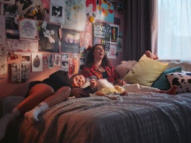 Two young white women sitting on a cosy bed, with lots of posters and art on the walls, laughing 