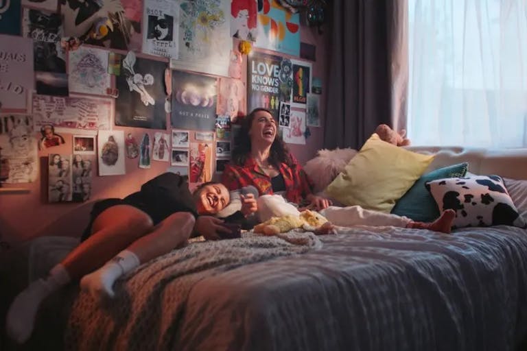 Two young white women sitting on a cosy bed, with lots of posters and art on the walls, laughing