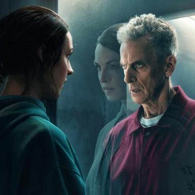 An older white man in a red jumpsuit looks menacingly through a two way mirror, a younger white woman stands on the other side 