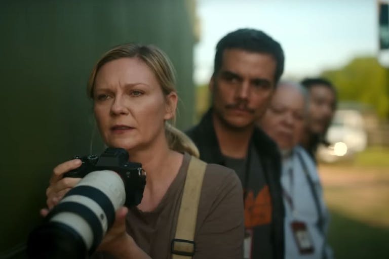 A white woman with a long lensed photo-journalist camera looking concerned, a line of men behind her