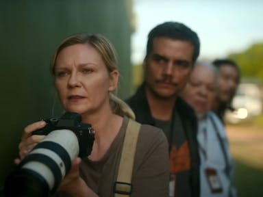 A white woman with a long lensed photo-journalist camera looking concerned, a line of men behind her