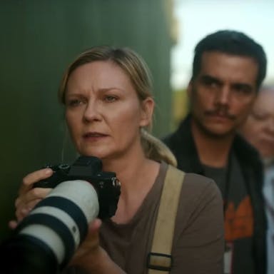 A white woman with a long lensed photo-journalist camera looking concerned, a line of men behind her