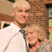 A tall blond male teenager standing next to his mum, leaning her head against his chest smiling