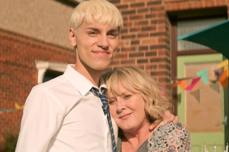 A tall blond male teenager standing next to his mum, leaning her head against his chest smiling 