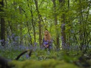 A young white woman with long blonde hair standing in a bright green forest with blue flowers