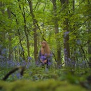A young white woman with long blonde hair standing in a bright green forest with blue flowers