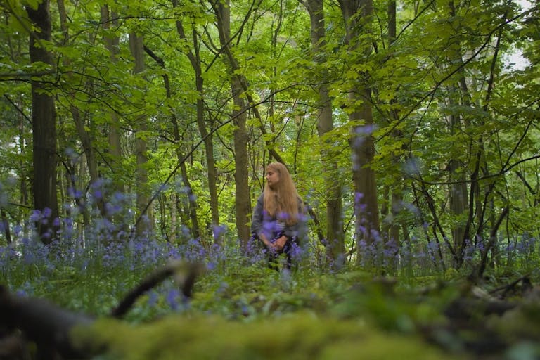 A young white woman with long blonde hair standing in a bright green forest with blue flowers