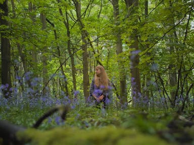 A young white woman with long blonde hair standing in a bright green forest with blue flowers