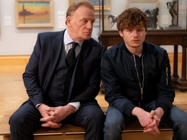 Young man and an older man in a suit sit on a bench next to each other in an art gallery