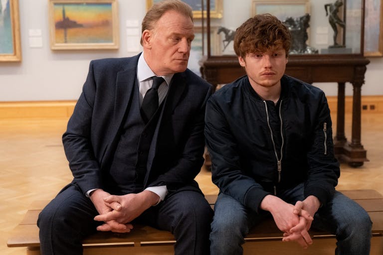 Young man and an older man in a suit sit on a bench next to each other in an art gallery