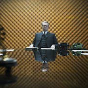 An older white man in a fine suit with silver hair combed back sits at a large stylish office table looking serious