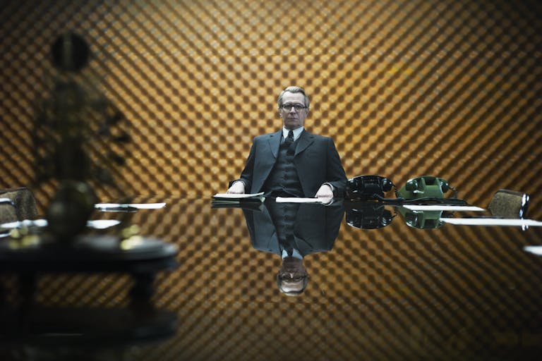 An older white man in a fine suit with silver hair combed back sits at a large stylish office table looking serious