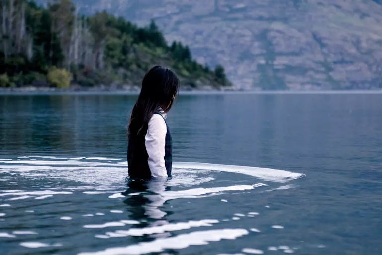 Young girl with dark hair, white shirt and black sweater vest, walking waist deep into a lake
