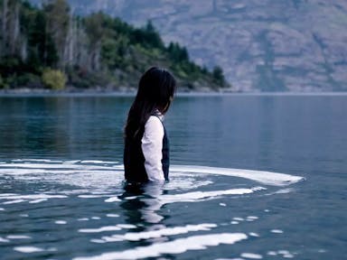 Young girl with dark hair, white shirt and black sweater vest, walking waist deep into a lake