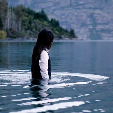 Young girl with dark hair, white shirt and black sweater vest, walking waist deep into a lake
