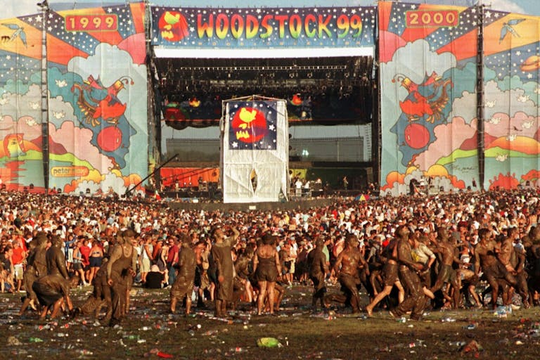 Large colourful stage with 'Woodstock 99' banner and a sea of muddy festival goers