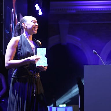 A Black woman in an elegant black dress on stage accepting a blue BIFA award