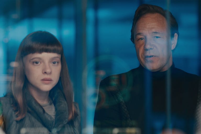 A young white woman and an older man stand behind reflected glass looking serious